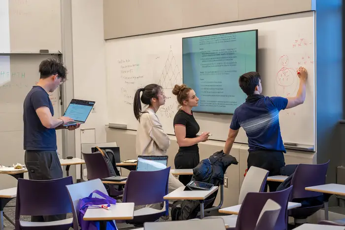 four students working at white board with one holding laptop