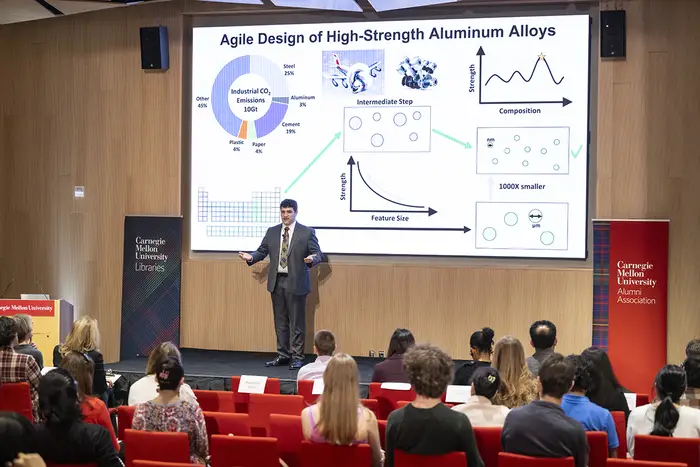 Benjamin Glaser on stage in front of seated audience with large screen presentation behind him