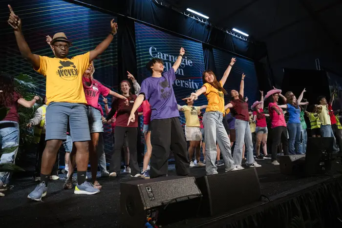 A large group of people with their arms raised wearing bright solid color shirts fill a stage in front of a large banner that says "Carnegie Mellon University".