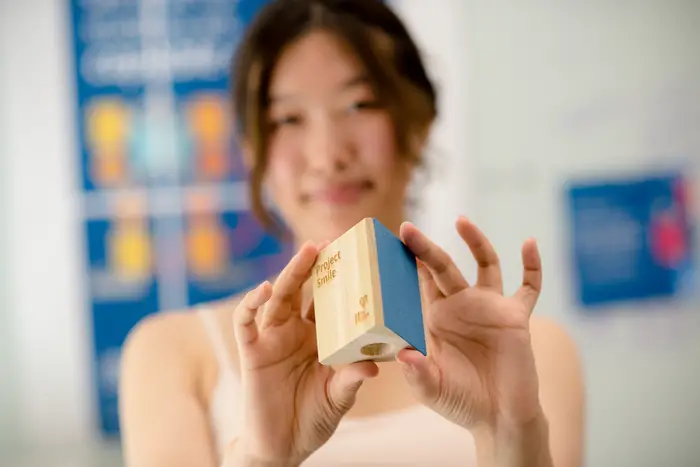 A woman out-of-focus in the background holds up a wooden block in-focus in the foreground.