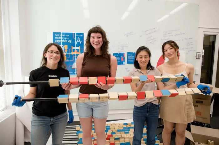 A group of four women hold up two horizontal metal rods that have wooden blocks threaded on to them with some of the blocks' sides painted blue or red.