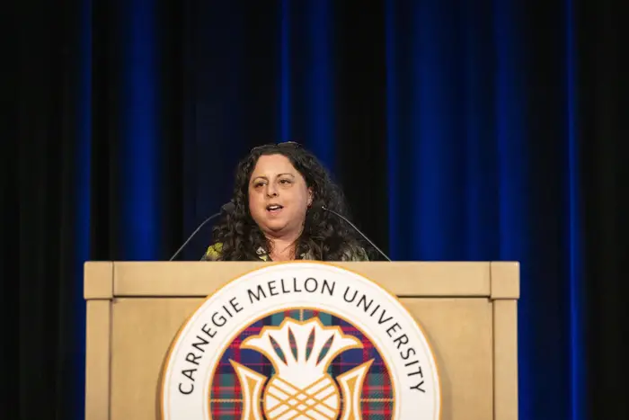 A white woman with shoulder-length dark hair speaks from behind a podium adorned with the Carnegie Mellon insignia