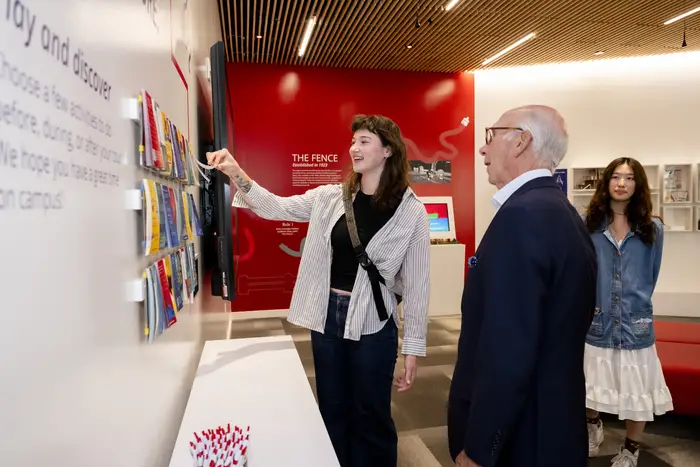 A woman lifts pages from a notepad secured with others along the wall in a display while a man and another woman look on.