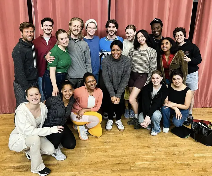 A group of about 15 students gather for a photo in front of a theater curtain with CMU alumna Jesmille Darbouze in the center.