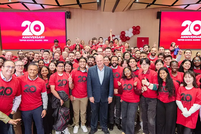 Large group of people all wearing the same red T-shirt, surrounded by a man in the business suit at the center. Large screens behind them are displying the same color red with white tex that says, "20th Anniversary - Tepper School naming 2024"