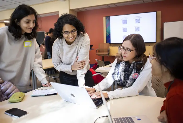 Two women stand next to a table talking with a woman seated at the table in the center behind a laptop with another seated woman next to her.