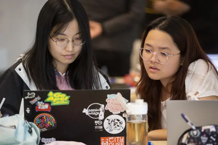 Two women sit at a table behind two laptops looking at one of the screens