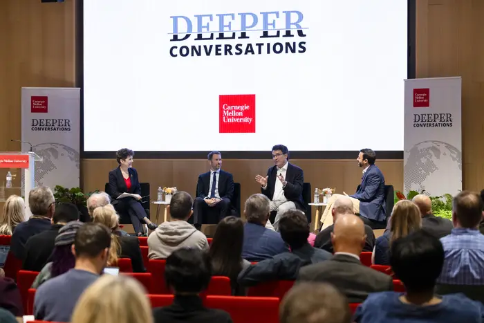 A group of four people on a stage sitting in chairs, in front of a screen that says "Deeper Conversations"