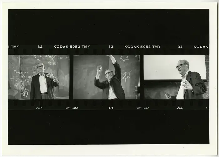 Filmstrip negatives with horizontal black bars across the top and bottom featuring three black-and-white photos of a man lecturing in front of a chalkboard with writing on it