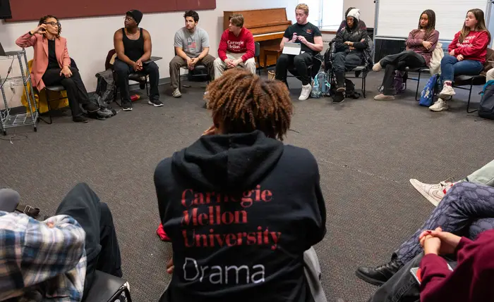 Students sit on chairs in a circle in a classroom while the professor, in the top left of the image, wearing a salmon-colored jacket talks and gestures.