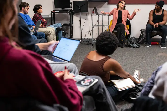 Students sit in a circle for class, with one holding open a laptop on their lap and another sitting on the floor, while the professor, a woman wearing a salmon-colored jacket, talks and gestures at the top of the image.