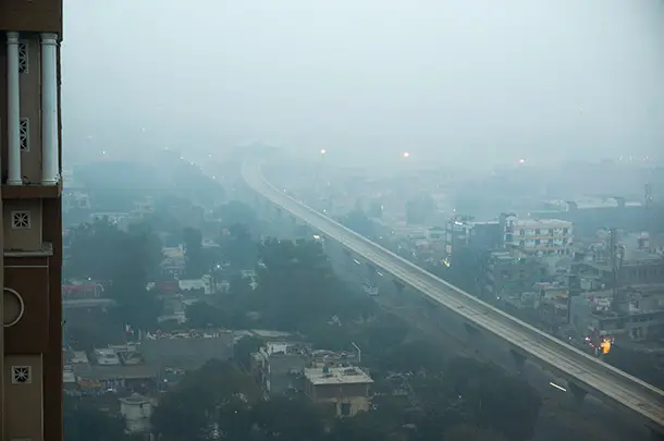 Buildings and a metro line viewed from above through thick fog. Even though it is morning, there is not much daylight, and bright streetlights and billboards are illuminated.