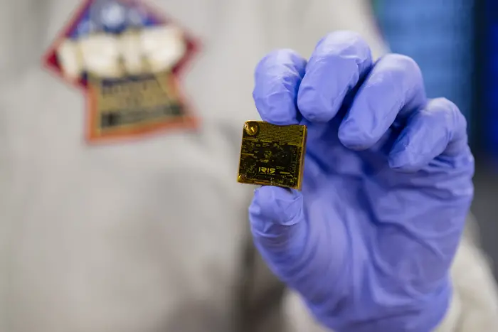 A hand in a blue glove holding a golden square object.