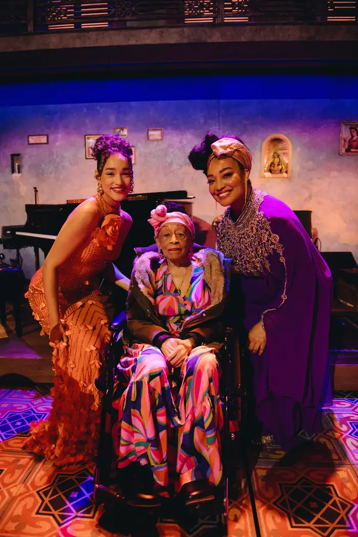 Two women in colorful dresses stand on either side of a third woman who is in a wheelchair.