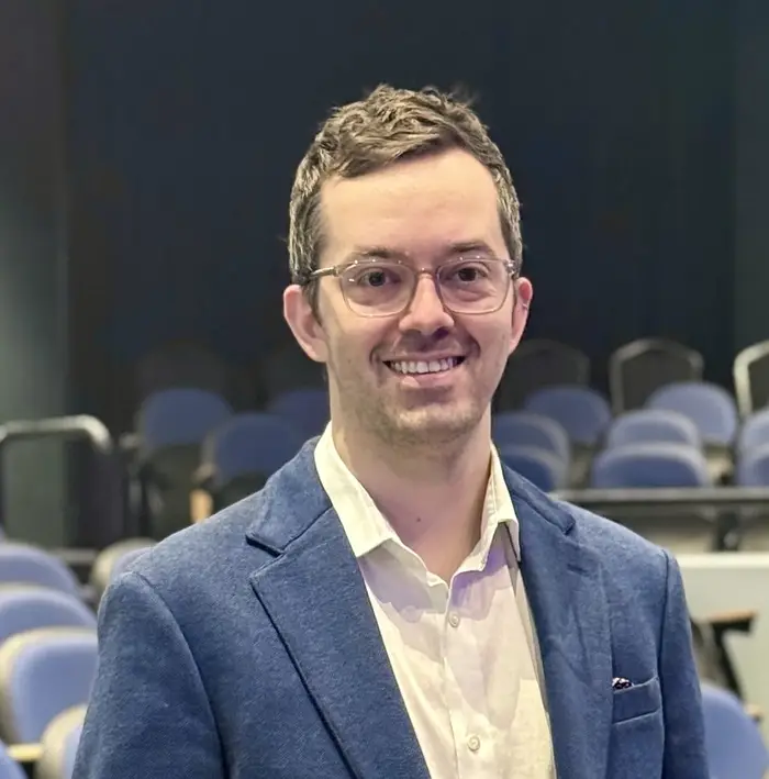 A head-and-shoulders crop of a man in a blue blazer and light yellow collared shirt wearing glasses smiles at the camera