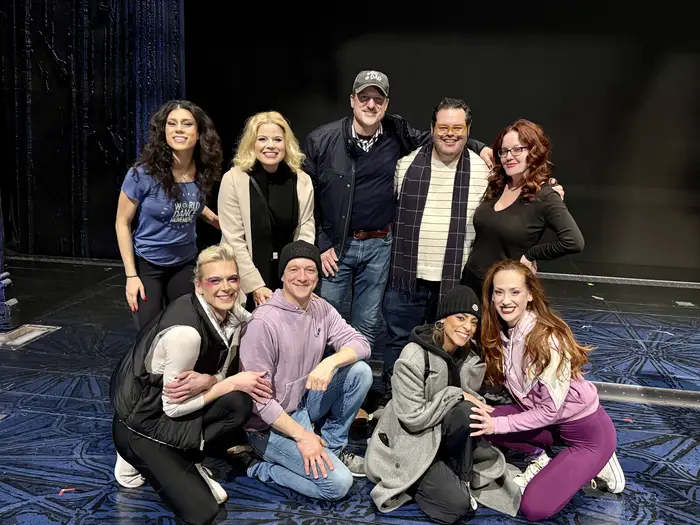 A group of nine people gather to pose and smile for a photo in two rows on a theater stage.