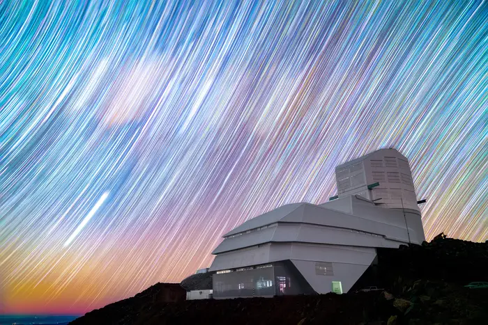 Star trails appear as colorful brushstrokes in the sky above a large observatory on a mountain.