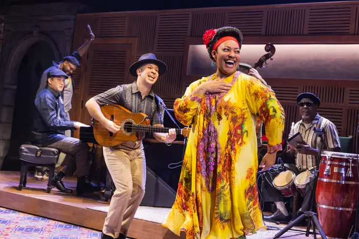 A woman in a yellow dress with flowers smiles while singing with four male musicians behind her including a man strumming a guitar and smiling, a man sitting and playing bongos, another man raising his hand and smiling next to a man seated and playing a piano.