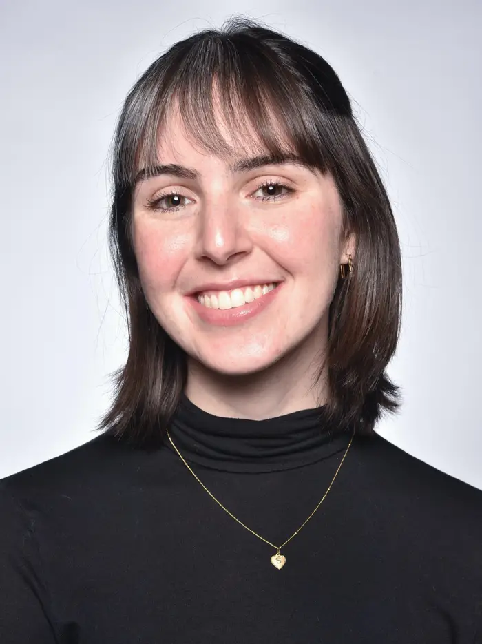A head-and-shoulders portrait of a smiling woman with shoulder length brown hair and bangs in a black turtleneck wearing a small gold chain necklace.