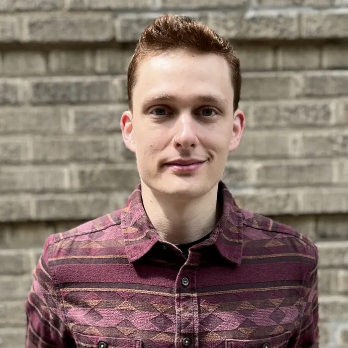 A head-and-shoulders crop of a photo of a man with short reddish hair wearing a red patterned collared shirt smiling at the camera