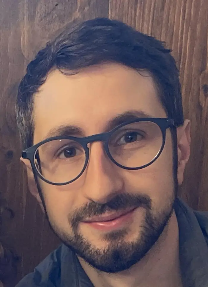 A close-cropped photo of the face of a man with beard, moustache, glasses and dark hair smiling at the camera.