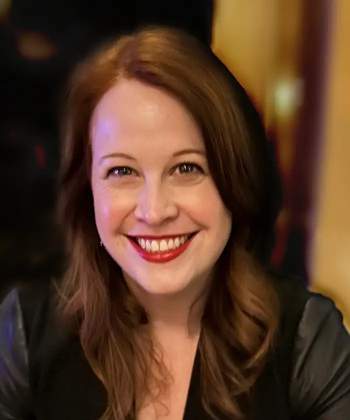 A head-and-shoulders portrait of a smiling woman with red lipstick and shoulder length brown hair.