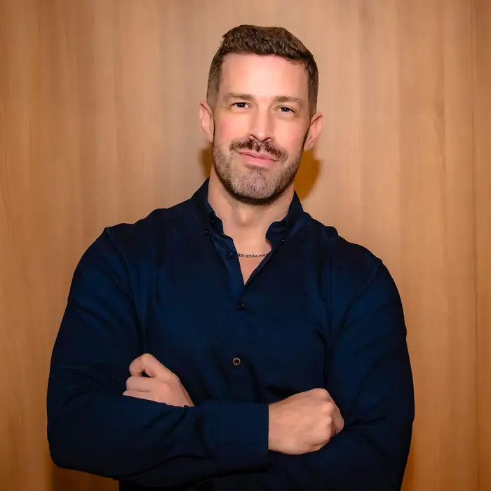 A waist-up photo of a brown-haired man with a moustache and five o'clock shadow wearing a black shirt stands with his arms crossed in front of him.