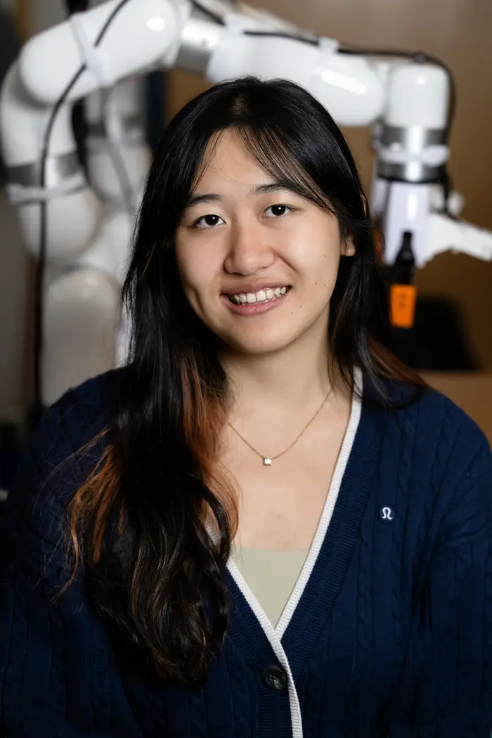 A woman with long dark hair wearing a dark blue V-neck sweater smiles at the camera with a white robotic arm attached that makes a "U" shape in the out-of-focus background over the woman's head.