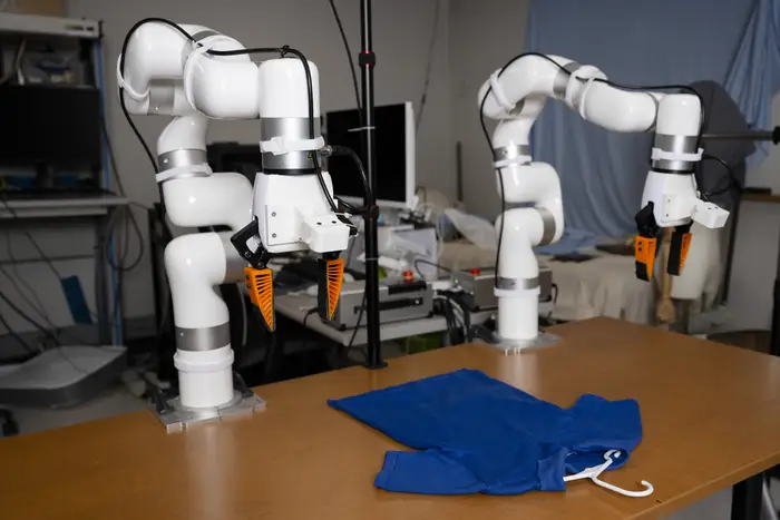 Two while robotic arms with silver bands at the joints and orange grabber tips perch in hook shapes over a wooden table where a blue shirt lays flat with a white hangar inside the collar.