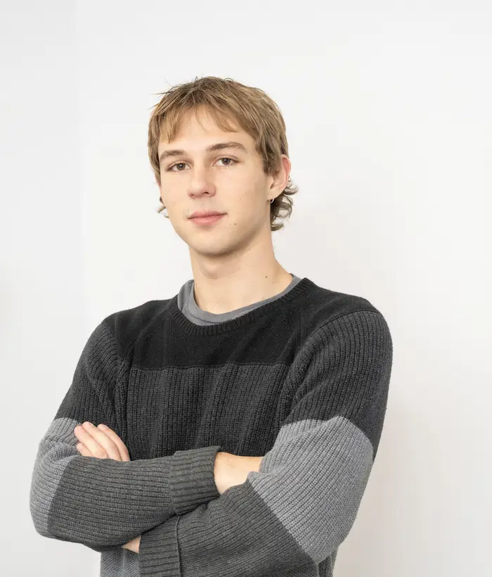 A young man with short blonde hair wearing a gray long-sleeved shirt stands for a portrait with his arms crossed, looking at the camera.