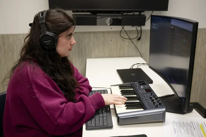 A woman with long black hair wearing headphones and a fuschia sweater plays a small piano keyboard attached to a computer as part of a music research study session.