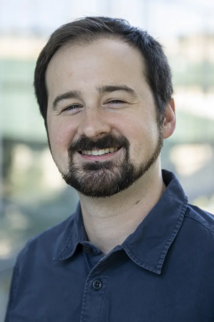 A man wearing a dark blue collared shirt who has brown hair and a brown beard and moustache smiles and looks at the camera