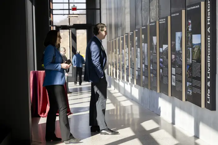 Two people look at a wall of flat panels comprising an exhibit of printed text and photos as the sun casts shadows through the windows behind them.