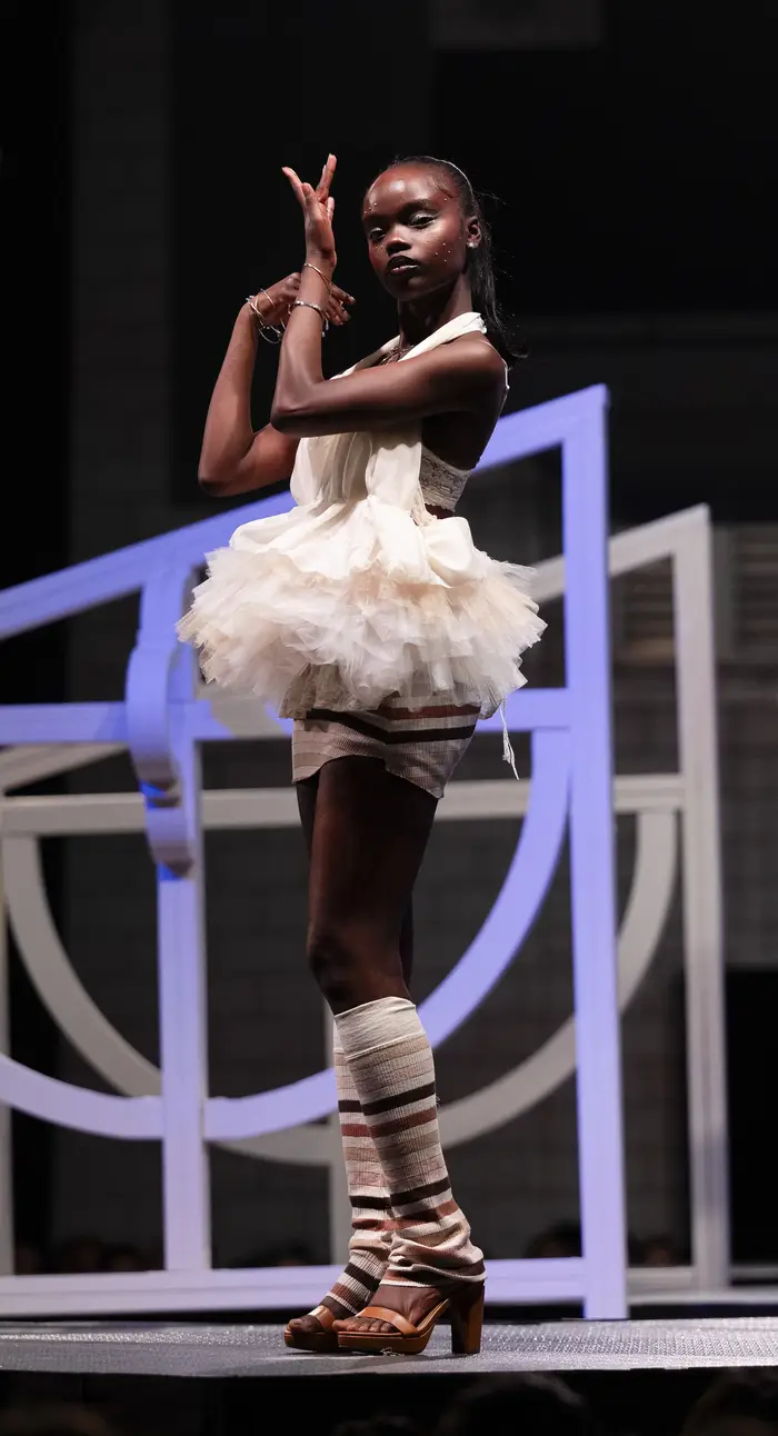 Model poses while wearing a short white babydoll dress with a poofy tulle tutu at her waist. She is also wearing high heels and leg warmers stripped white, mauve, and navy