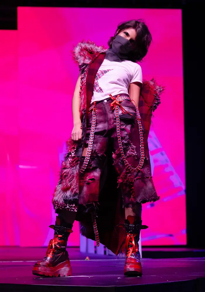 Model is wearing a white t-shirt with an embroidered red plaid star on its side, and red pants made up of a patchwork of leather, denim, embroidery, and fur, with calf-high black and red boots