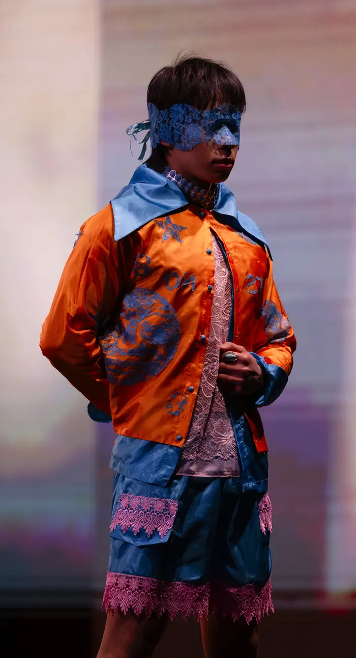 Male model wearing a blue lace eye mask and an orange shirt with a wide sky blue collar and sky blue embroidery along the torso. This is complemented by sky blue shorts with pink lace edging along the bottom