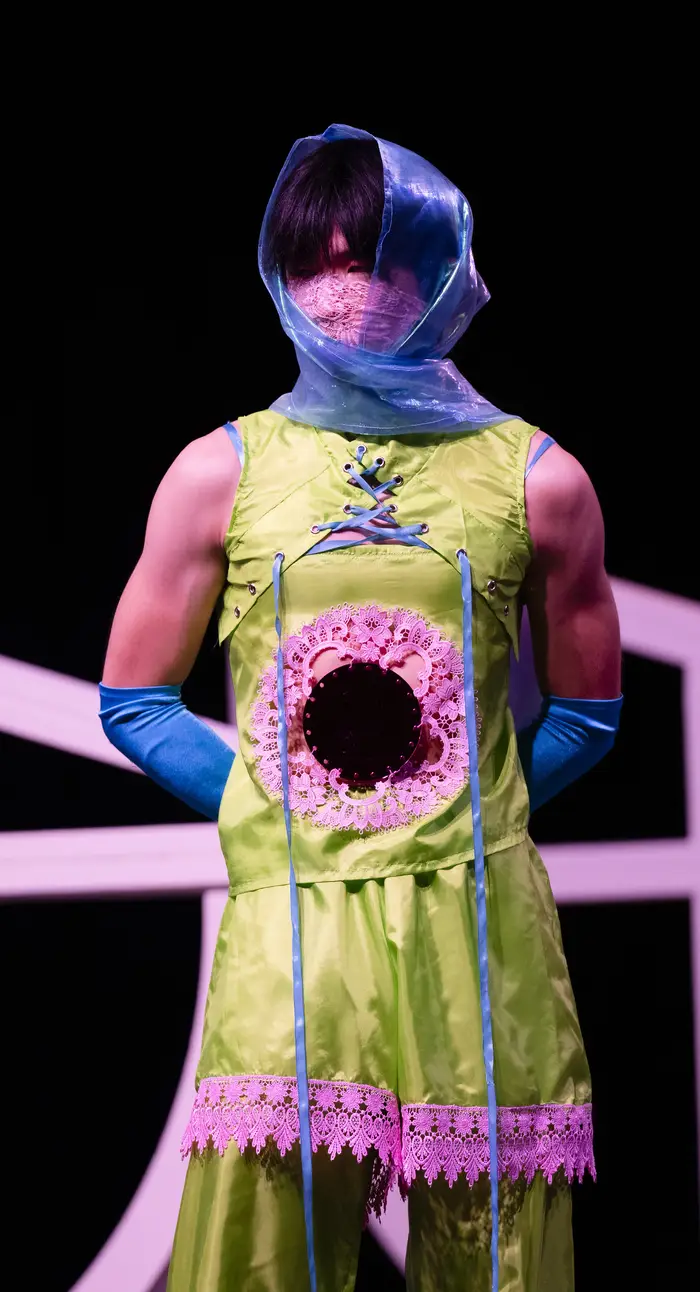 Male model wearing a chartreuse green tunic and pants, with hot pink lace edging on the tunic's bottom and middle, as well as an indigo headscarf and gloves