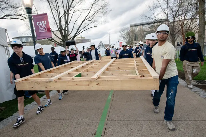 Students carry a large wooden construction across CMU campus while wearing hardhats. 