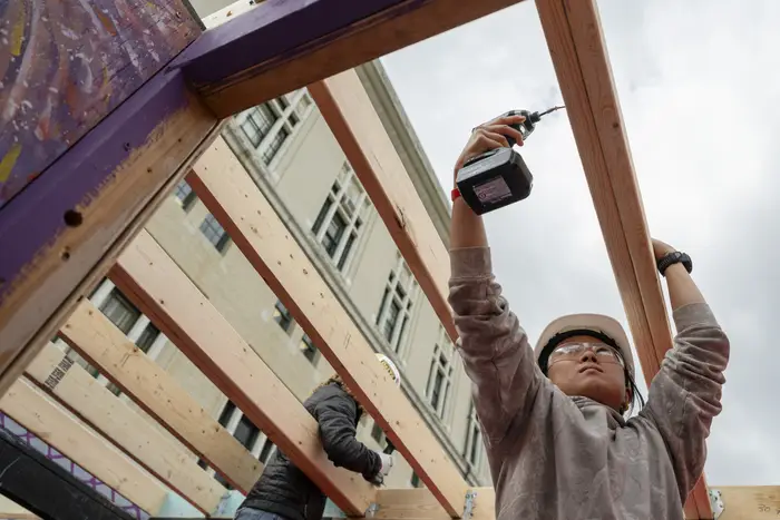 A woman in a hardhat drills a wooden frame. 