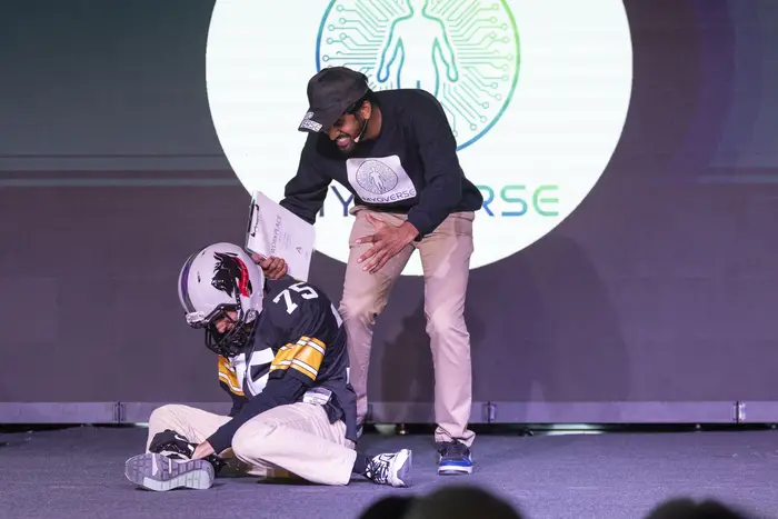 A person on stage in football pads and a helmet sits on the ground while another person talks to them. 