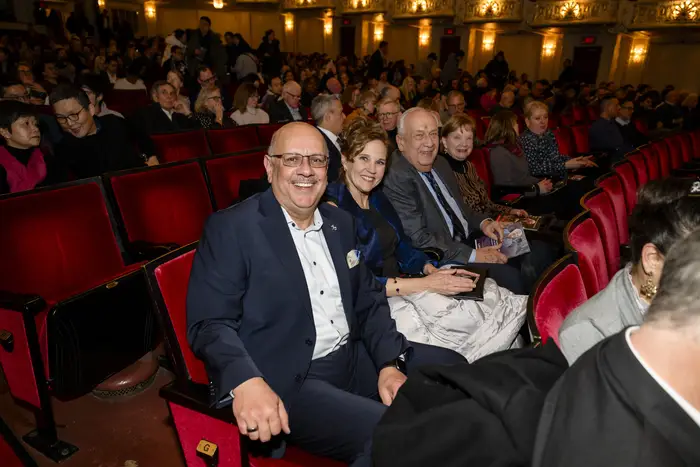 A row of people sit in red theater seats with other audience members in the seats around them.