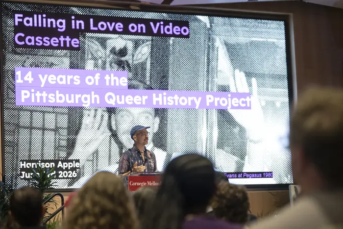 A person stands behind a podium in front of a screen that says "Falling in Love on Video Casette: 14 years of the Pittsburgh Queer History Project" with heads of seated audience members in the foreground.