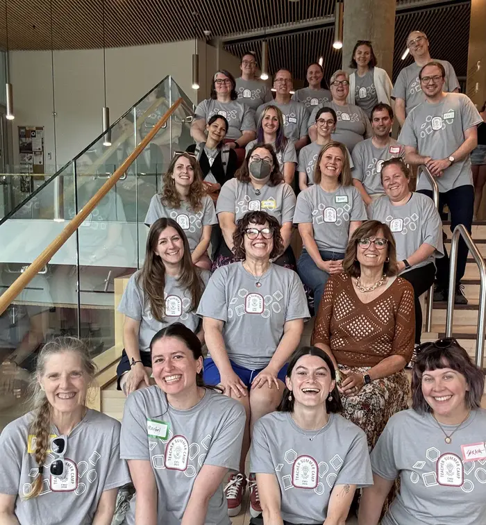 A large group of about 30 people all wearing the same gray T-shirt sit on stairs and smile at the camera with one woman with glasses in the second row from the front wearing a brown top.