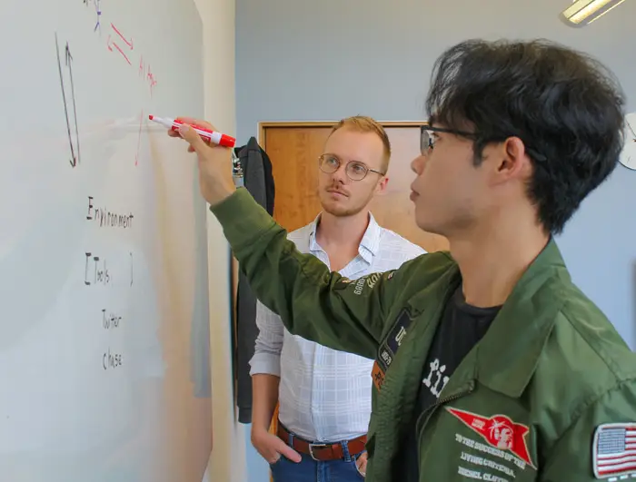 A man with black hair and glasses wearing a dark green jacket and black t-shirt writes on a whiteboard while another man with blonde hair and glasses looks on from the background.