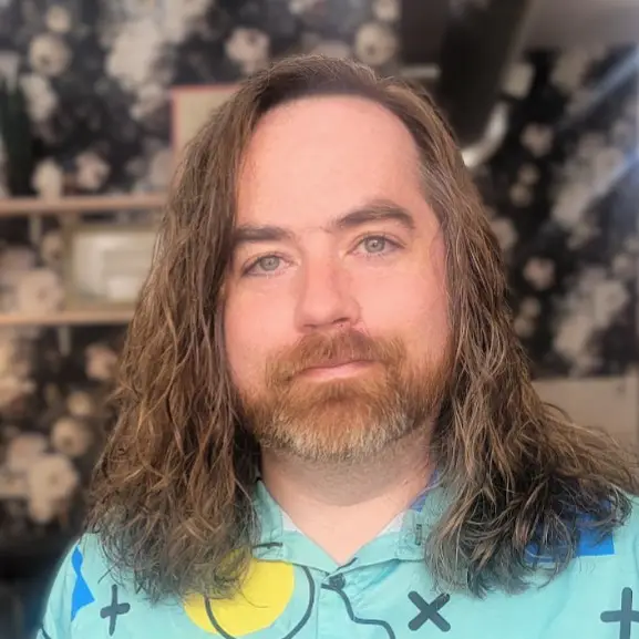 A man with shoulder-length brown hair, a beard and moustache poses for a headshot while wearing a aquamarine collared shirt.