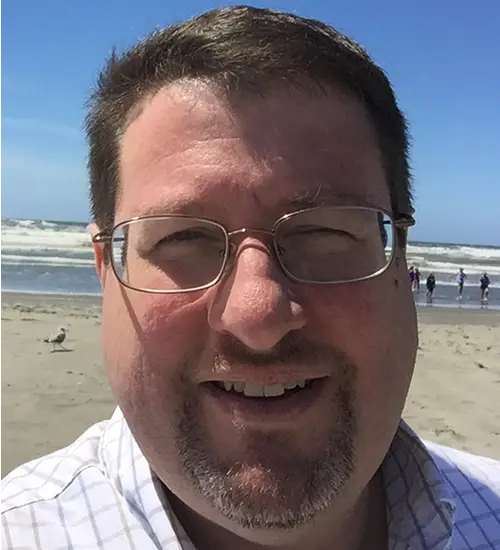 A man wearing glasses and a collared shirt smiles in a headshot in front of a seashore.