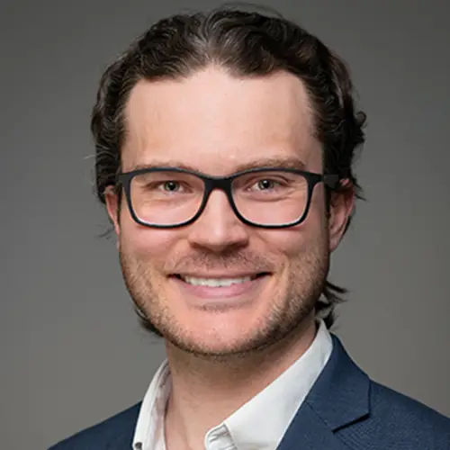 A man with dark hair wearing glasses, a white collared shirt and a dark blue jacket smiles looking into the camera.