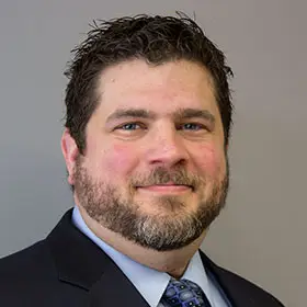 A man with dark hair, a beard and moustache wearing a dark suit jacket, light blue collared shirt and dark blue tie smiles at the camera.