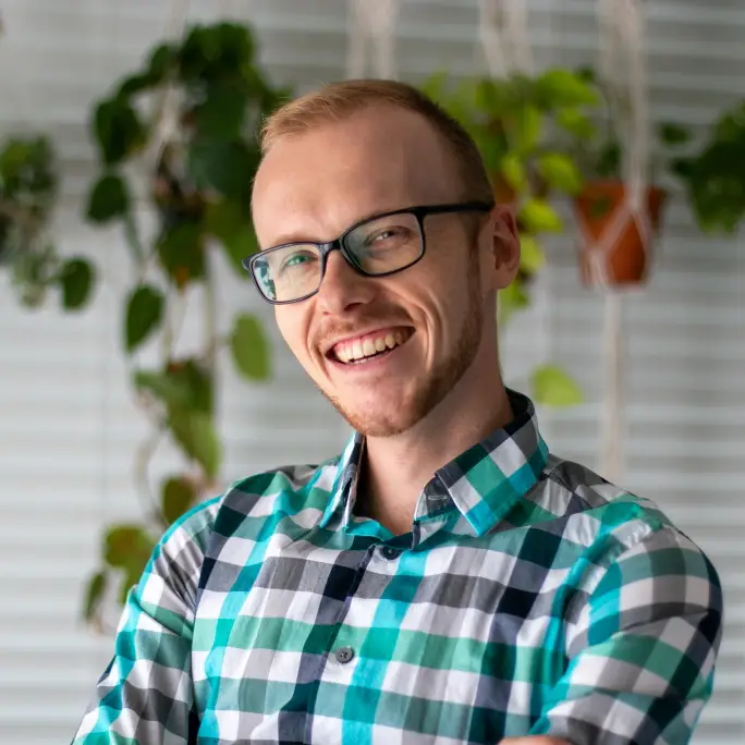 A man with short blonde hair wearing glasses and a blue and gray checkered shirt standing in front of a hanging plant and white window blinds smiles for the camera.