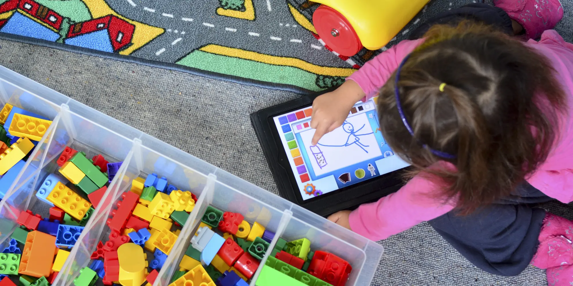 a student sits on the floor and plays with a drawing application on a tablet next to bins of colorful blocks